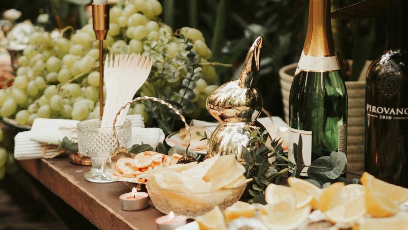 A table full of food (notably white grapes) and Champagne. There are candles and a plate of lemon